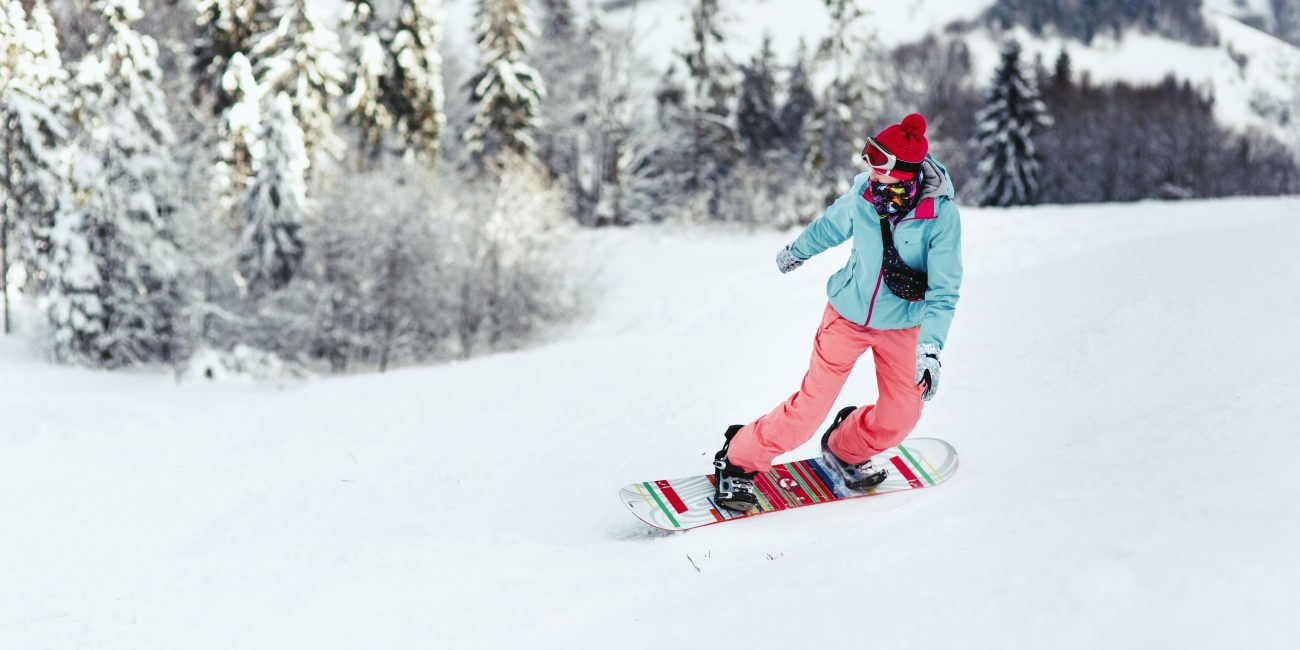 woman in ski suit looks over her shoulder going down the hill on her snowboard